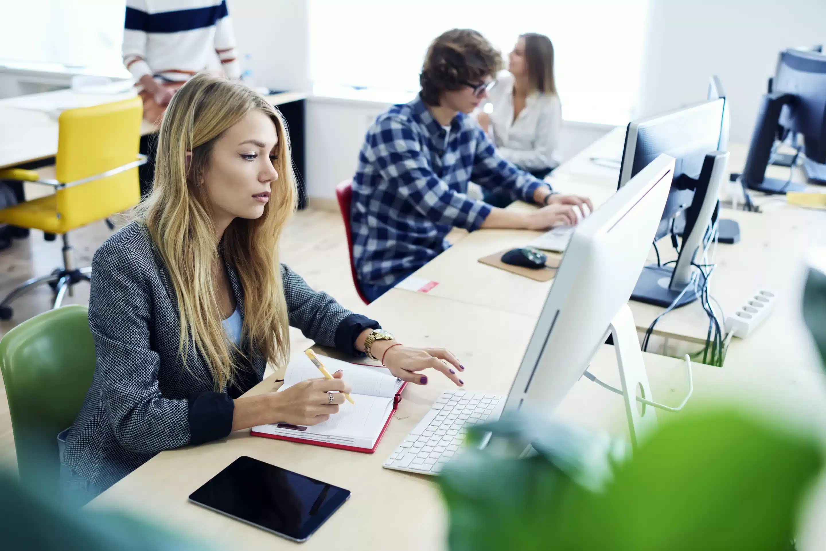 a woman sitting at a desk with a computer and a man in the background