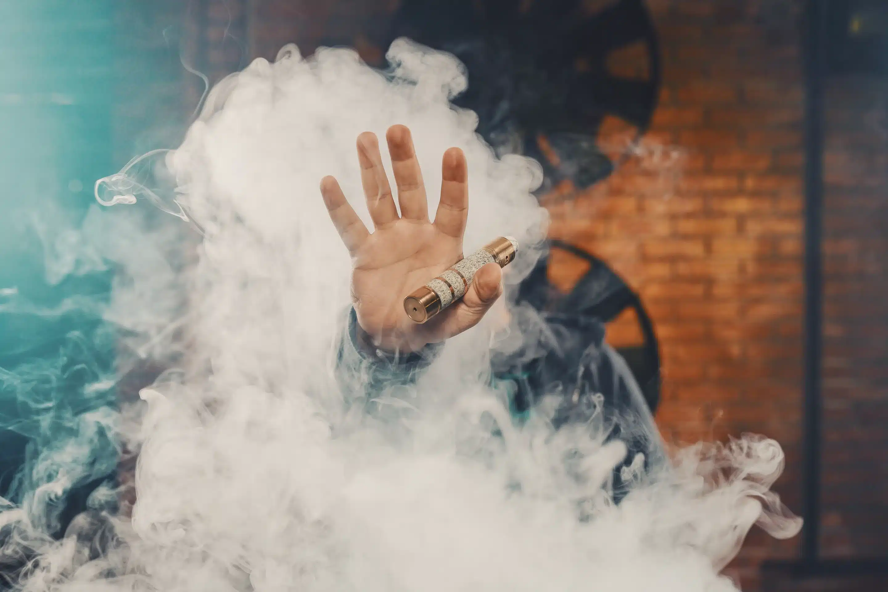 Vaping man wearing a hat, holding up a mod, obscured behind a cloud of vapor. 