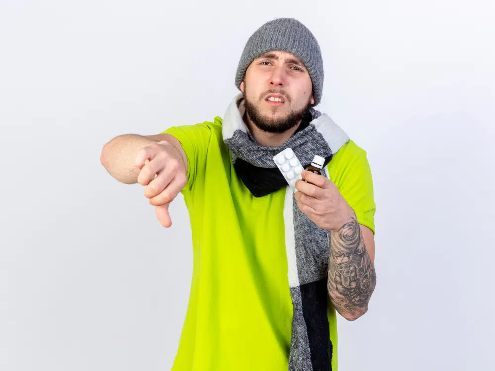 Upset sick young man in winter clothes giving thumbs down while holding medicine