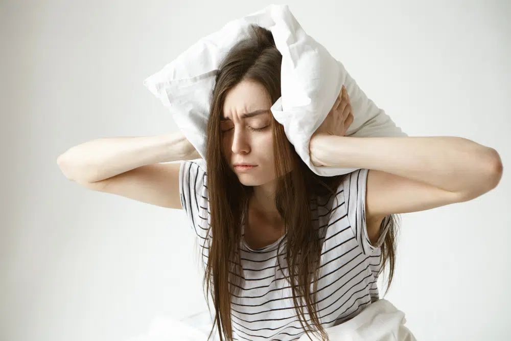 Stressed young brunette woman with anxiety wearing striped pajamas, sitting anxiously on bed
